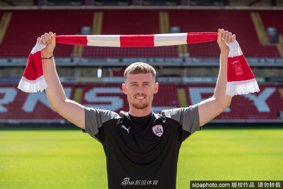 罗比·库迪手持巴恩斯利球衣，在俱乐部标志前自信微笑，眼中闪烁着对新挑战的憧憬