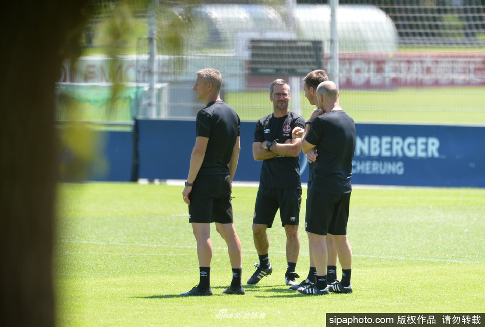 远景下的训练场，一群穿着明亮橙色背心的球员在翠绿的草坪上整齐奔跑，仿佛烈日下跳动不息的火焰。