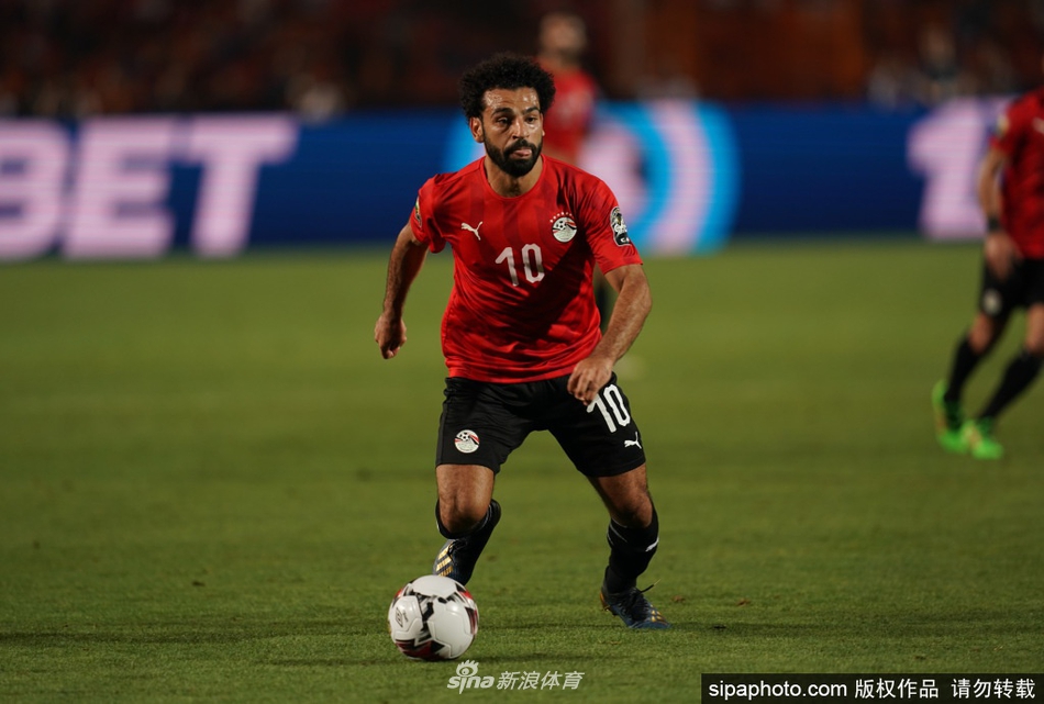 身披埃及国家队战袍的萨拉赫在绿茵场上全速冲刺，身后是飘扬的埃及国旗，象征着他为国家荣誉而战的决心