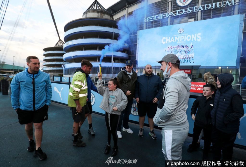 一名年轻的曼城小球迷骑在父亲的肩头，他用力挥舞着一条巨大的曼城围巾，稚嫩的脸庞上洋溢着无法掩饰的骄傲与喜悦，身后是同样沉浸在狂欢中的球迷人群。