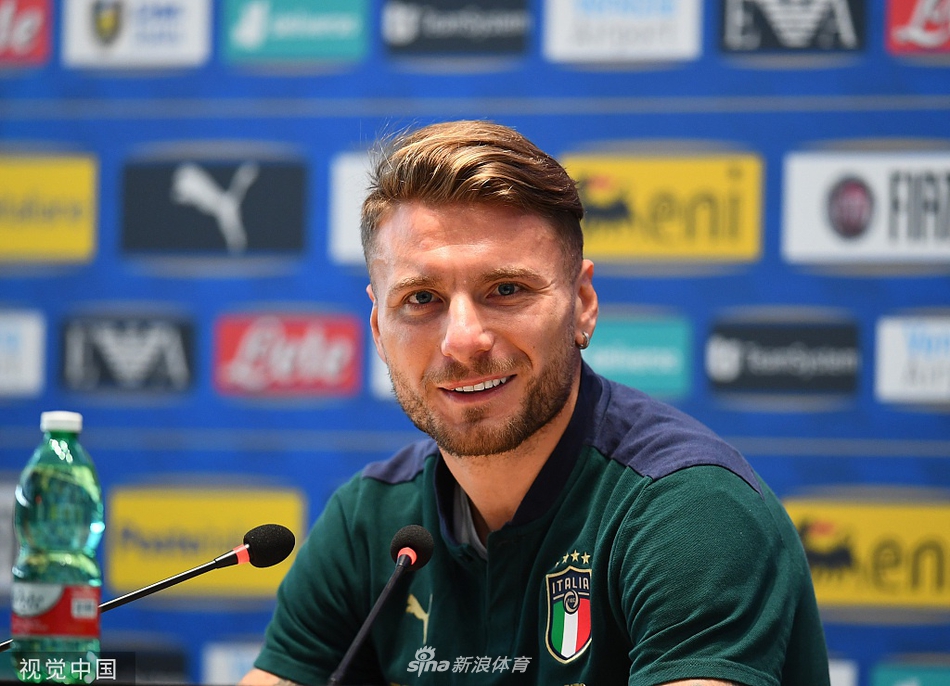 因莫比莱神情专注地倾听记者提问，他微微前倾的身体姿态显示出他对每一个问题的重视，身后的意大利队徽在灯光下格外醒目。