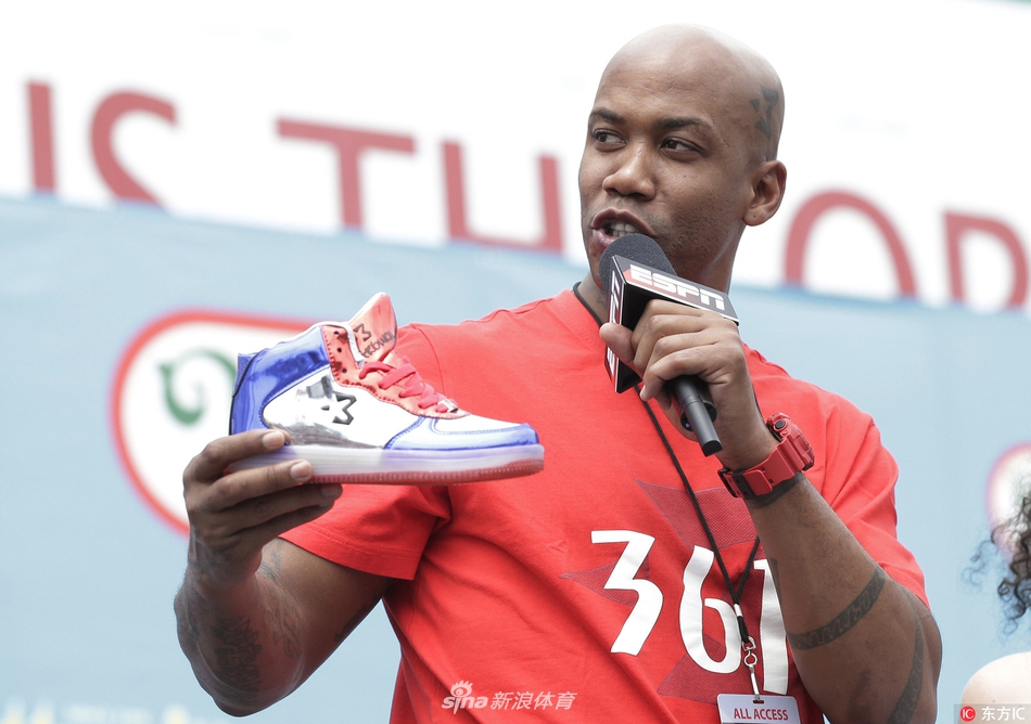 马布里现身纽约科尼岛热狗大赛现场，身穿深色紧身运动衣搭配个人品牌球裤，面带微笑望向镜头，背景是熙熙攘攘的围观人群和美国国旗装饰。