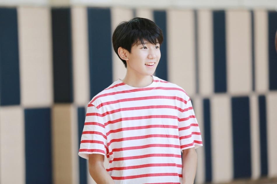 王源在球场中央舒展身体如飞鸟展翅，红色条纹衫下摆扬起，黑色运动裤与背景的绿茵形成鲜明对比，定格青春最美的剪影