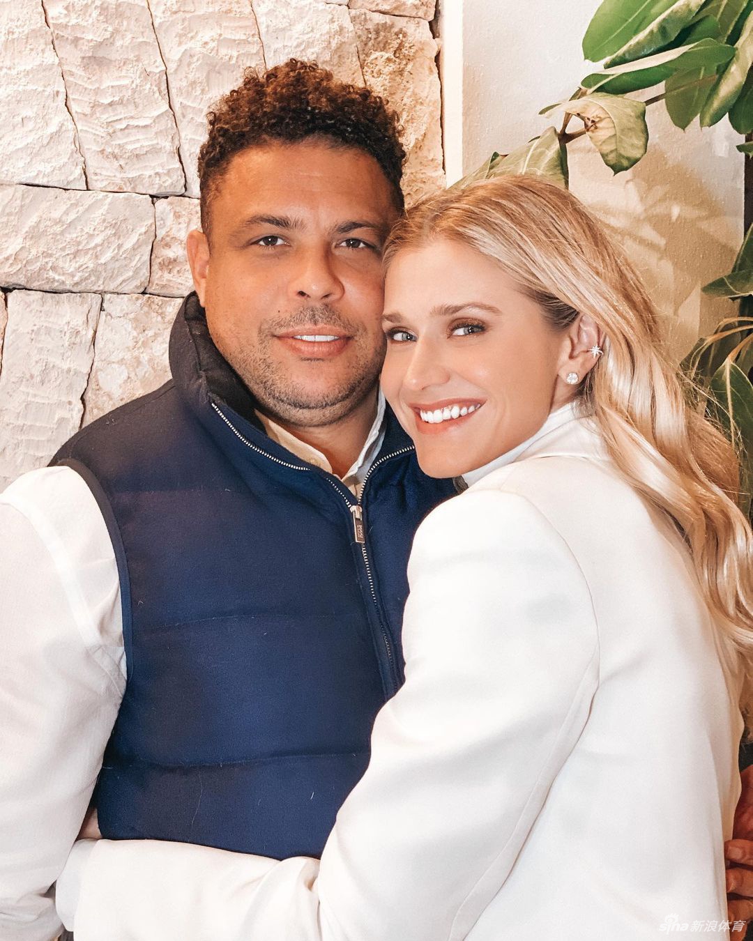 赛琳娜穿着白色蕾丝连衣裙，优雅地坐在海边餐厅的椅子上，她双手轻轻交叠，似乎在为腹中的宝宝祈祷，脸上带着恬静而期待的神情