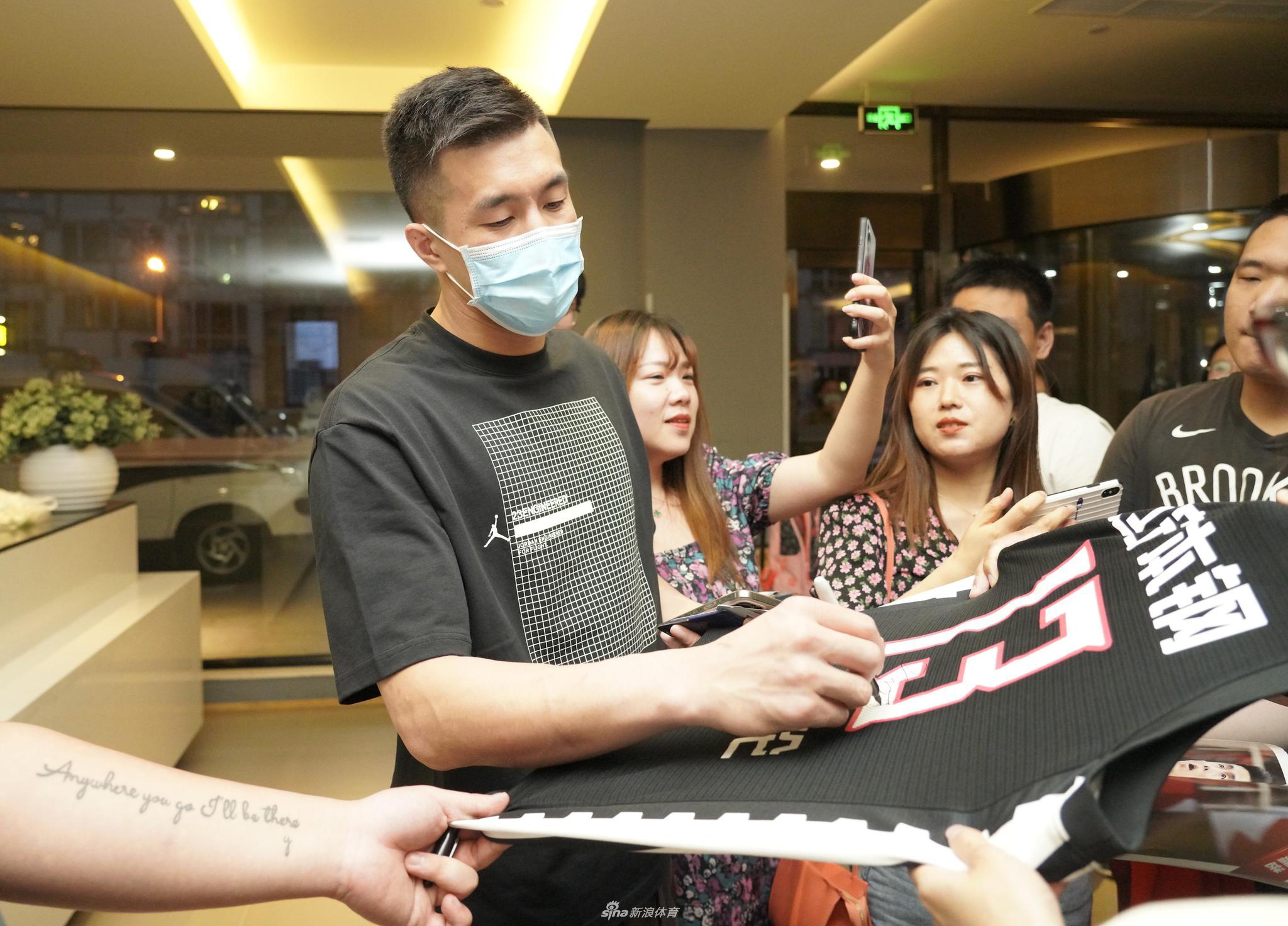 郭艾伦手拿签字笔，全神贯注地在物品上签名，旁边的女粉丝手持照片耐心等待，背景是北京集训驻地的玻璃门。