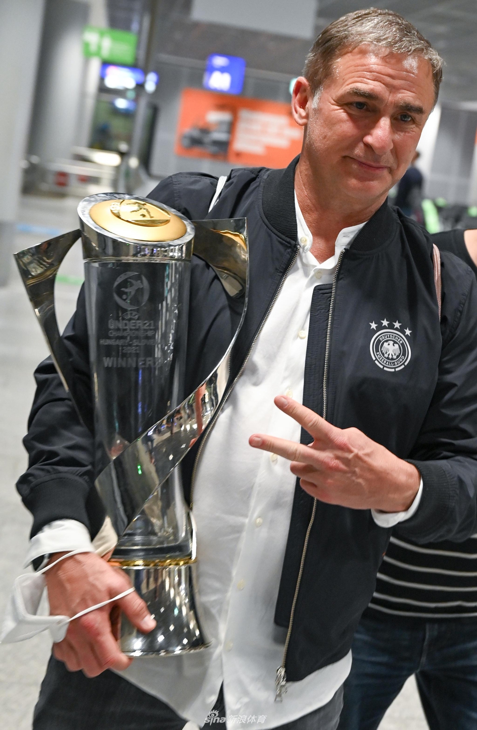 冠军旗帜下的少年英雄，德国U21队员在法兰克福机场骄傲地举起欧青赛冠军奖杯，脸上洋溢着难以掩饰的喜悦与自豪