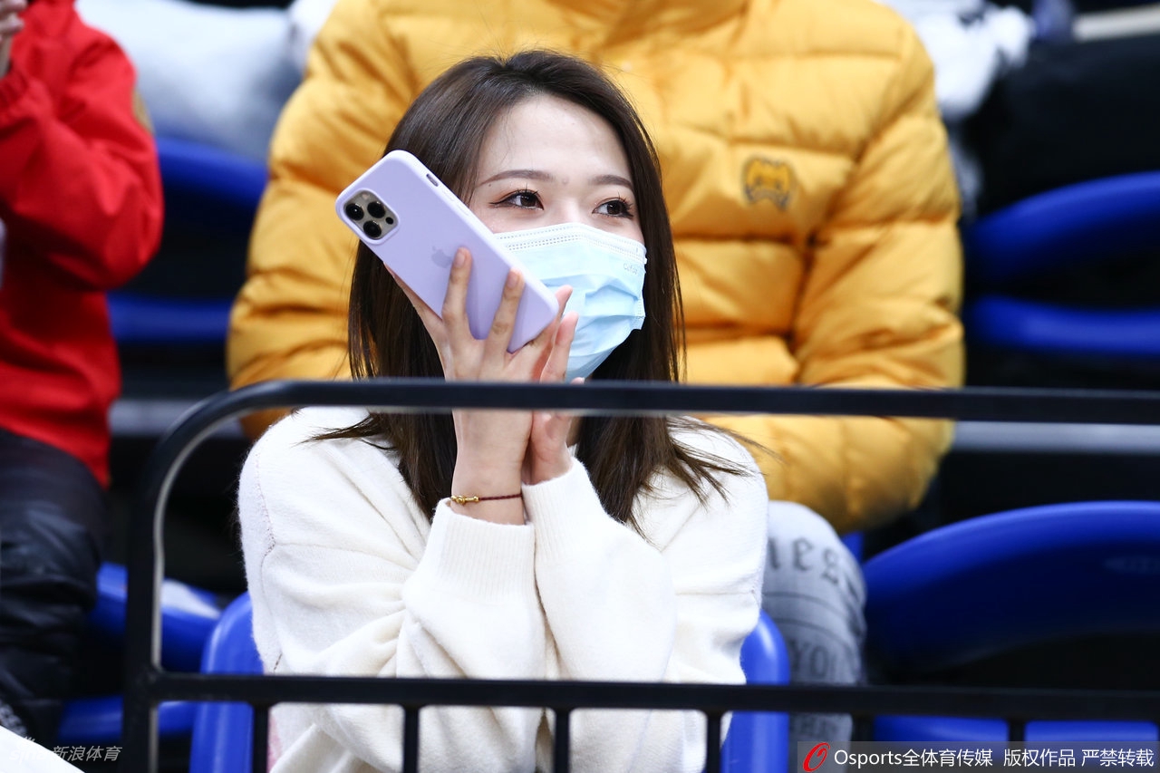 一位长发披肩的靓丽女球迷，面戴口罩难掩眉眼笑意，正专注地用手机记录赛场上的精彩瞬间。