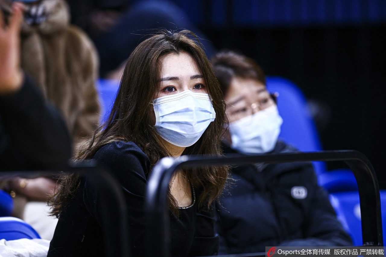 一位身穿深色卫衣的温柔女球迷，手拿应援物，安静地坐在看台上，眼神清澈，默默守护着心中的主队。