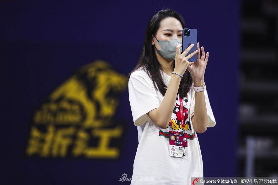 记者转过身来，目光追随着飞向篮筐的篮球，右手轻轻抬起放在身前，姿态优雅从容，背景是模糊的观众席。