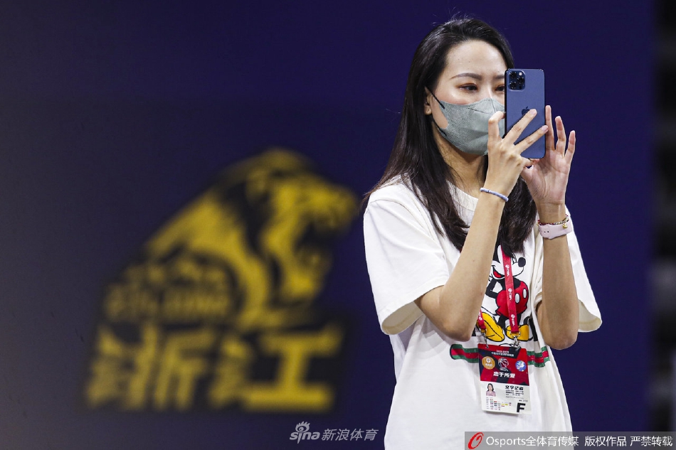 美女记者侧身而立，脸上浮现出微微的笑意，似乎为场上的精彩进球而赞叹，柔和的光线勾勒出她精致的面部轮廓。