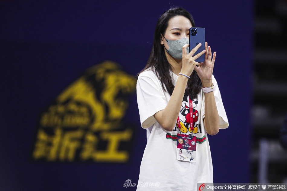 身姿挺拔的美女记者立于篮板后方，她身着米色风衣，神情专注地望向赛场，仿佛与紧张的战局融为一体。