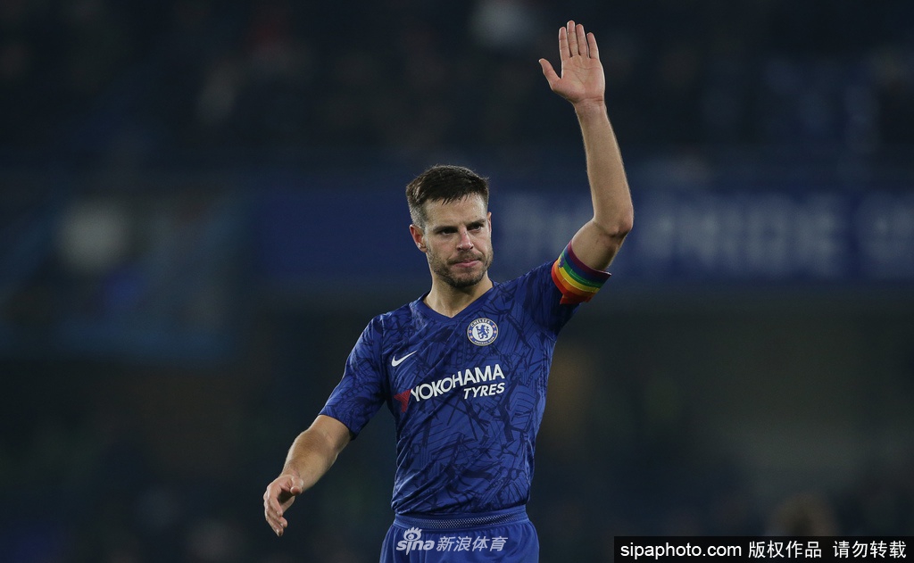 镜头聚焦在球场边的角旗区，蓝色的切尔西队徽在微风中轻轻摆动，背景是空荡荡的看台和上方巨大的电子显示屏，整个画面透出一种肃穆而又坚韧的力量。