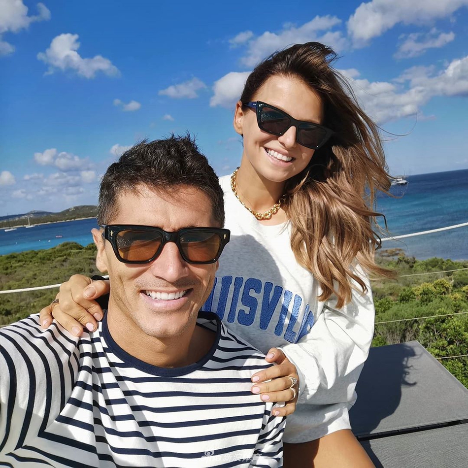 莱万多夫斯基赤裸上身，展现出堪比顶级模特的健硕肌肉和完美线条，他微笑看向镜头，与身穿白色连衣裙的妻子相视而笑，画面充满活力与美感。