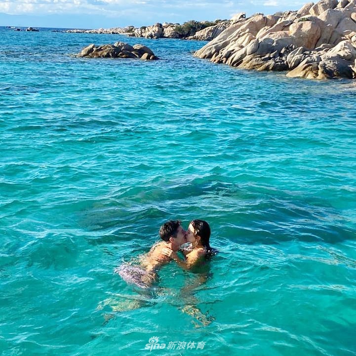 莱万与妻子在度假地亲密依偎，他带着墨镜的表情轻松惬意，妻子的笑容温暖明媚，两人背后是迷人的海岸风光，构成一幅完美的画卷。