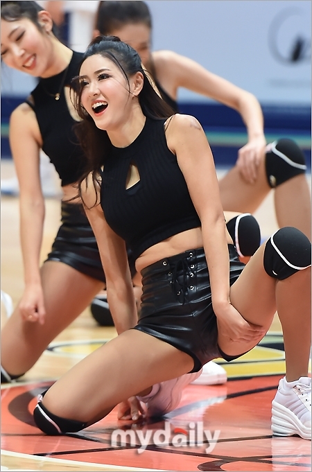 一位啦啦队员侧身对镜头，乌黑的长发随着舞动飘扬，她身穿黑色皮短裤，上身是活力四射的白色露脐上衣，完美的身材曲线在聚光灯下展露无遗，仿佛定格了最动人的舞蹈瞬间