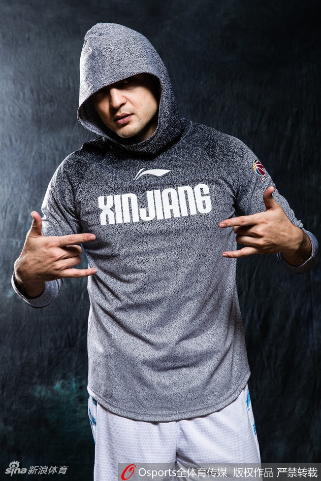 西热力江坐在椅子上身体前倾,双手交叉,身着时尚运动装,表情严肃而富有攻击性,如同场上指挥交通的组织后卫