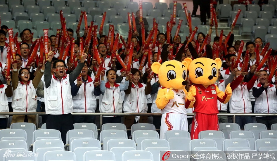 吉祥物站在看台台阶上向二楼的观众抛洒小礼物,它仰着头用力挥舞着手中的纪念品,灯光照亮了它身上红色的球衣,上方无数双手臂高高举起,仿佛一片欢呼的森林