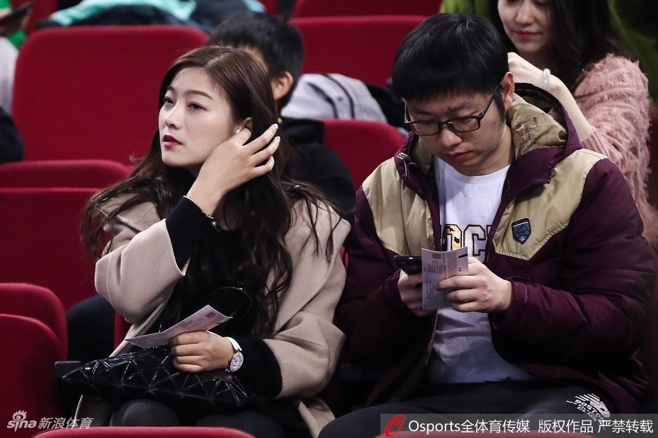 一位长发美女站在看台通道处回眸，眼神中带着一丝笑意。她身穿简约的深色外套，身后是涌动的人群和赛场明亮的灯光，整个画面充满了动态的美感，仿佛定格了比赛间隙的一个迷人瞬间。