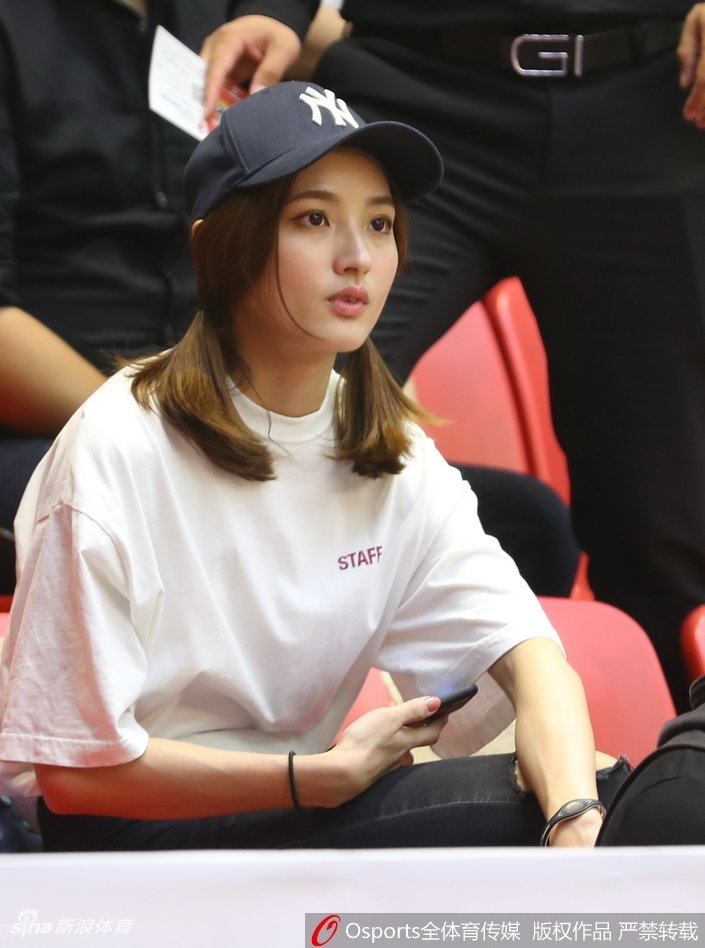 一位气质清冷的女球迷在场边安静地观看比赛，柔和的灯光洒在她的侧脸上，与身后喧嚣的庆祝人群形成鲜明对比