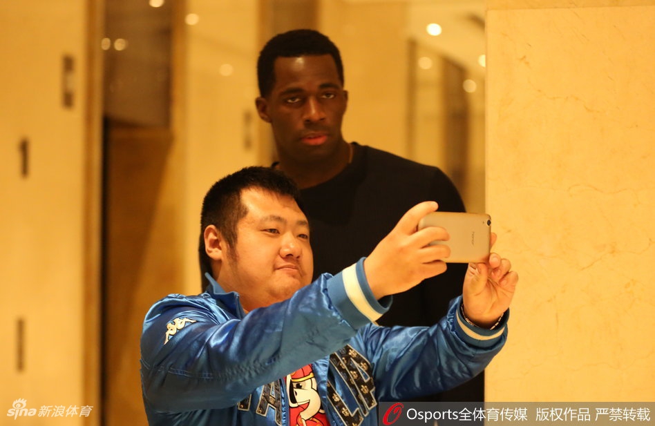 酒店门口，一位满头白发的老球迷紧紧握着主教练郭士强的手，激动得说不出话来。郭士强指导弯腰倾听，眼神中充满了感激与敬意。背景中，球迷们挥舞着围巾，将这一刻的温情永远定格。