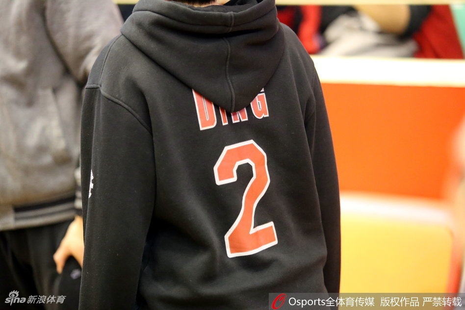 看台上的球迷们集体起立，人群中可见多件丁彦雨航主题的文化衫，整个看台被橙色的助威色覆盖，气氛热烈