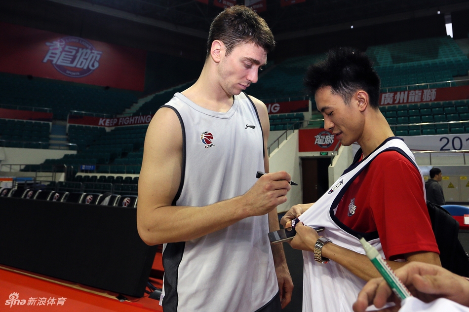 广州男篮开放日的趣味对抗环节,一名球员高高跃起试图封盖队友的投篮,旁边其他的队友和场边的球迷都被这一瞬间逗乐,气氛欢快。