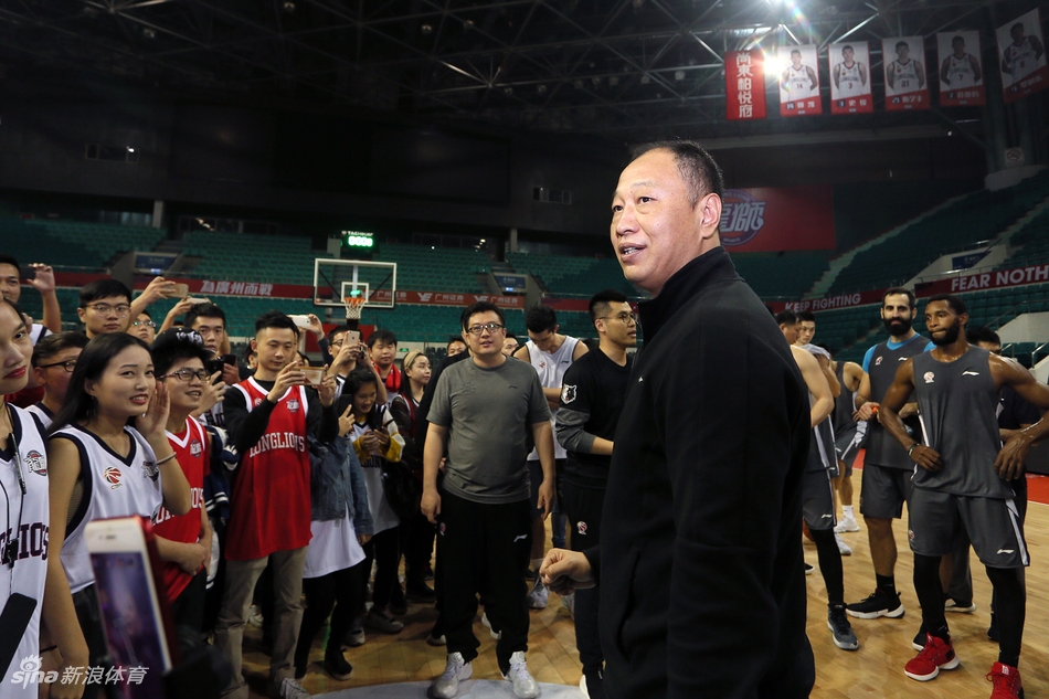 广州龙狮男篮开放日现场,球迷们围坐在场边,专注地看着场地中央正在进行的训练,背景是明亮的篮球馆和悬挂的旗帜。