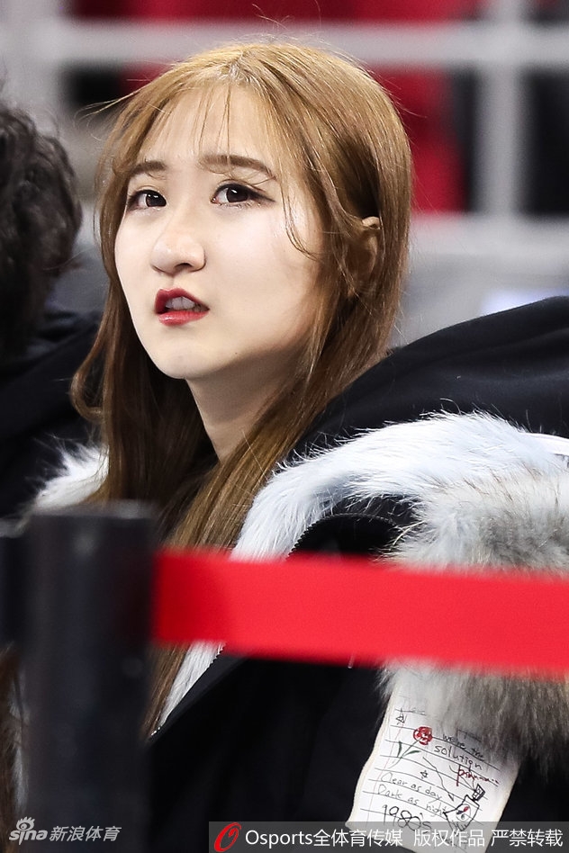 一位身着浅灰绒衣的温婉女球迷坐在看台上，眼神专注而柔和，仿佛整个球场的喧嚣都与她无关，她的眼里只有那一道橘红色的弧线。