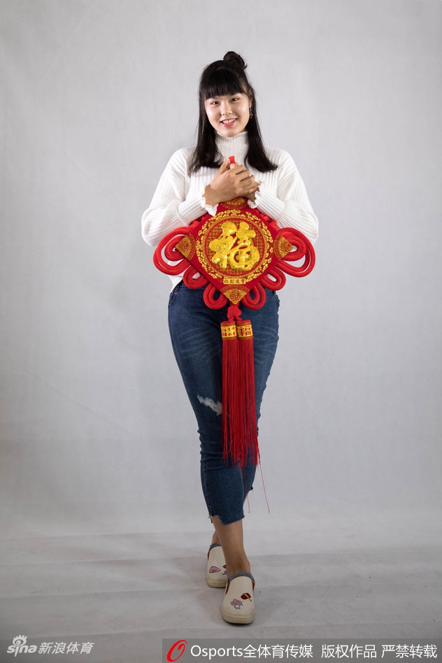 黄思静手持一张精致的金色福字卡，对着镜头俏皮地眨眼，展现出赛场之外活泼可爱的一面，背景中隐约可见其他队友正在相互整理着装，气氛温馨和谐。