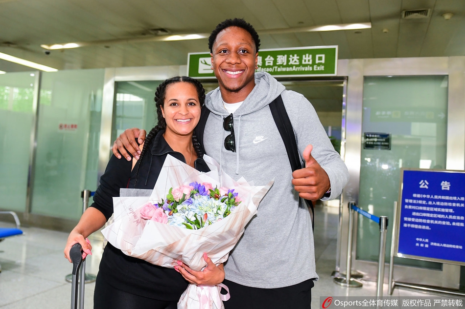 卡南的女友怀抱鲜花面对镜头展露甜美笑容，卡南则侧身看向远处，两人的穿搭休闲时尚，成为机场一道亮丽风景。