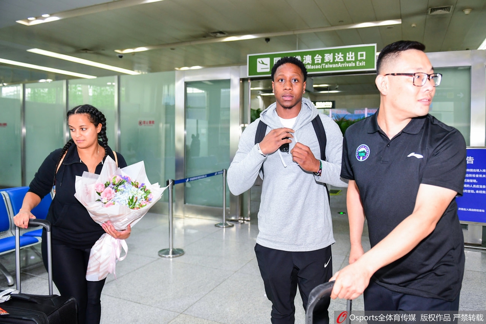 在机场一角，卡南与友人亲切交谈，女友安静地站在一旁注视，卡南做出手势，似乎正在讲述旅途见闻，气氛融洽。