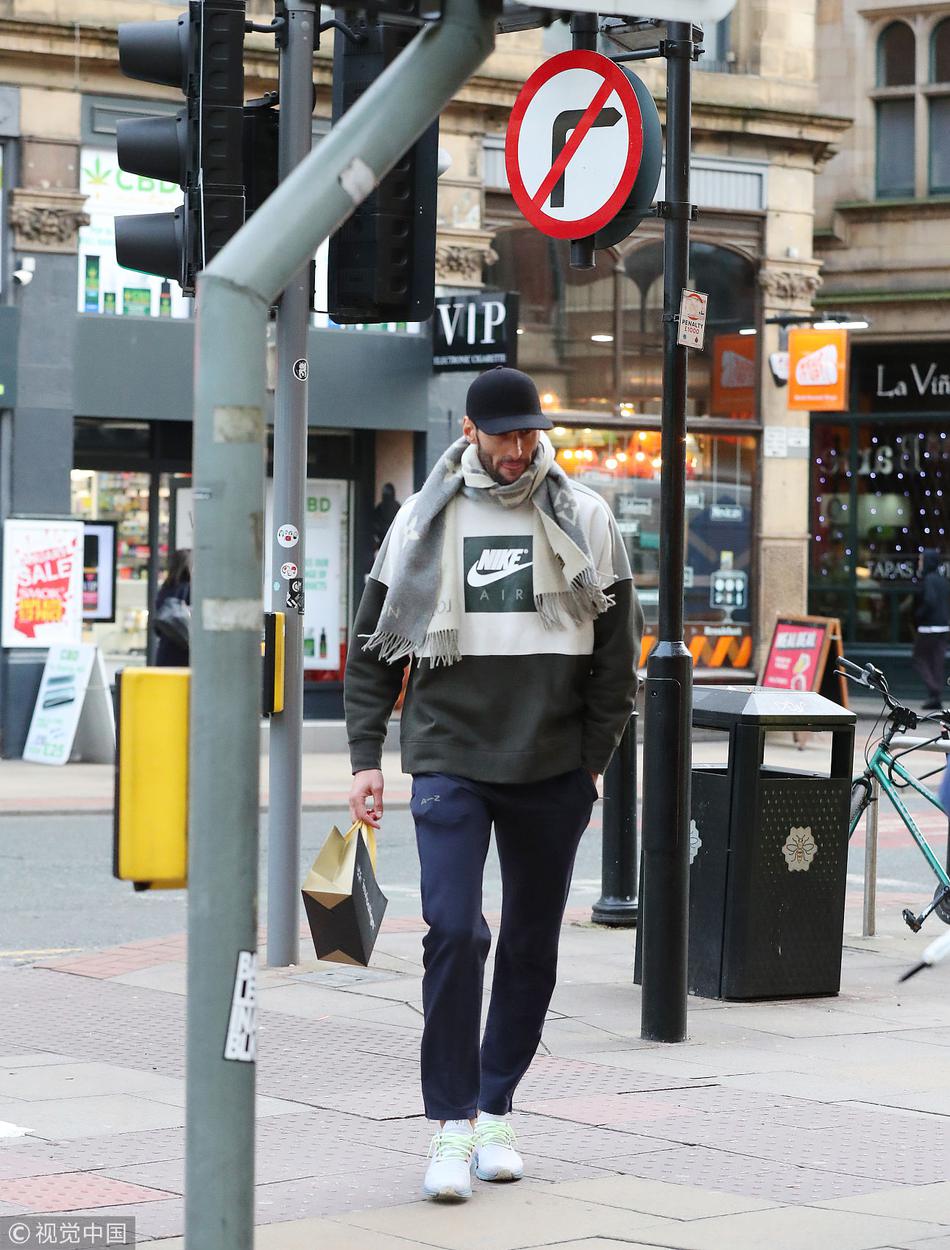 在人流稀疏的街道拐角，费莱尼独自一人面对斑马线。身后巨大的商业广告牌色彩斑斓，与他深色的穿着形成强烈对比。他仿佛站在命运的路口，即将做出改变职业生涯轨迹的重要决定，那孤独的背影与周围冰冷的城市景观融为一体。