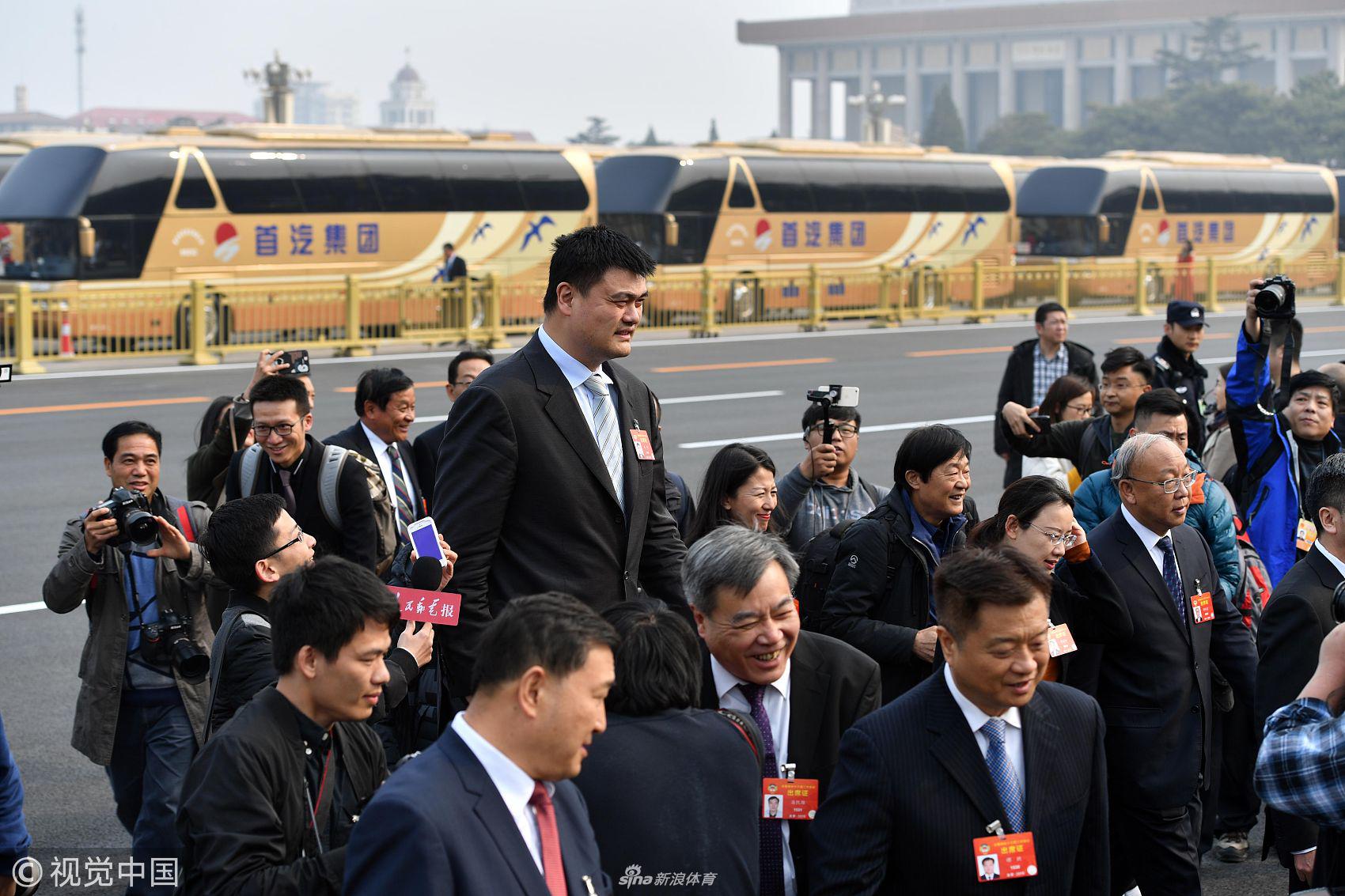 人民大会堂内，姚明在小组会议中认真倾听同组委员发言，巨大的身形在红色座椅上显得格外突出，但他的表情却显得谦逊而专注，手中紧握着笔随时准备记录。