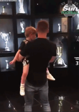 在尤文图斯的更衣室里，拉姆塞将儿子抱在怀里，指向一个衣柜，仿佛在说：'看，这是爸爸的新位置！'小家伙则好奇地伸手想触摸衣柜上的队徽。