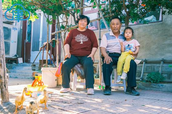 村民周景西夫妇和小孙女在院子里葡萄树下合影，平时顽皮的小孙女面对镜头还有些腼腆。