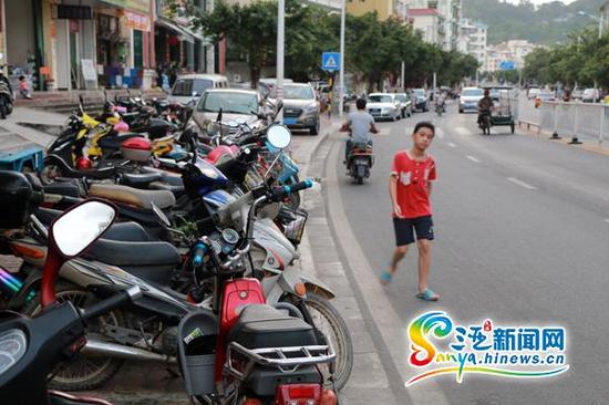 在三亚市南边海路南海山庄附近，人行道被电动车、摩托车、私家车占据，一位少年只能在机动车道上通行。少年后方的不远处，3辆私家车就停放在机动车道上。(三亚新闻网记者 邓松摄)