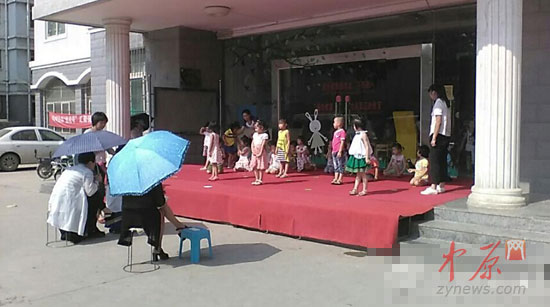 幼儿园小朋友在园门前的空地上排练，据该幼儿园工作人员称，该园园长当时就撑伞坐在下面。