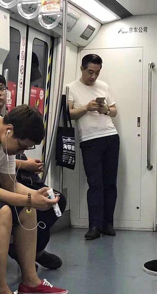 张召忠身着白色T恤，斜倚在地铁车厢内玩手机。  