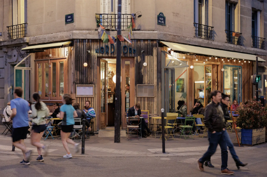 巴黎某咖啡馆外，行人与慢跑者络绎不绝，露天座位上零散坐着一些用餐的顾客。