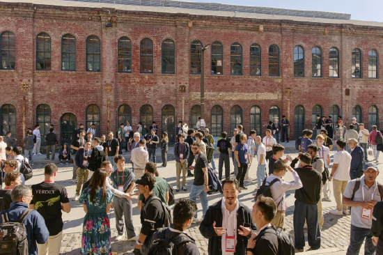大批年轻人聚集在历史红砖建筑前的鹅卵石广场，大家衣着休闲、佩戴工牌，三五成群交流社交
