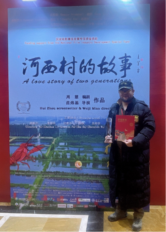 出品人、总导演苗炜基手捧荣誉奖杯和荣誉证书在影片《河西村的故事》海报前