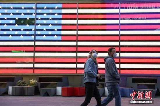 资料图：当地时间4月12日，两位戴口罩的市民走过纽约时代广场美国国旗灯箱。中新社记者 廖攀 摄