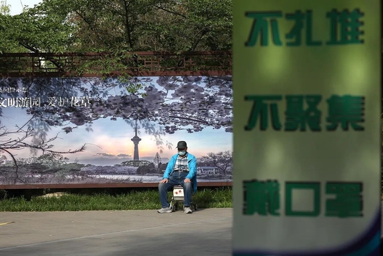 2022年5月18日，北京，公园内一位市民在“不扎堆、不聚集、戴口罩”的标语旁休息。