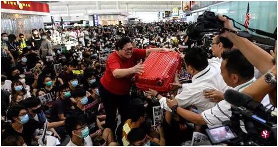 ▲8月13日下午，一名准备飞往新加坡的女子被示威者阻挡去路，高举红色行李箱，跨过数百示威者（图源：法新社）