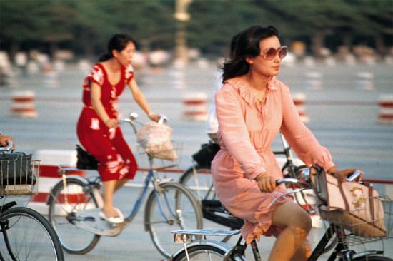 1985年夏，北京街头骑车的女青年。烫大波浪头、戴墨镜、穿粉色镶花边裙装，这位女性绝对是引领时尚的弄潮儿，她身后的红裙女士也毫不逊色。1984年，电影《街上流行红裙子》就反映了那一时期的社会风潮。摄影|李晓斌