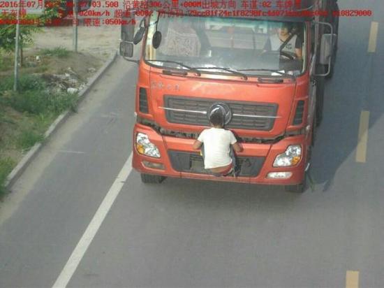 图为一男子挂在大货车前挡住车牌。图
