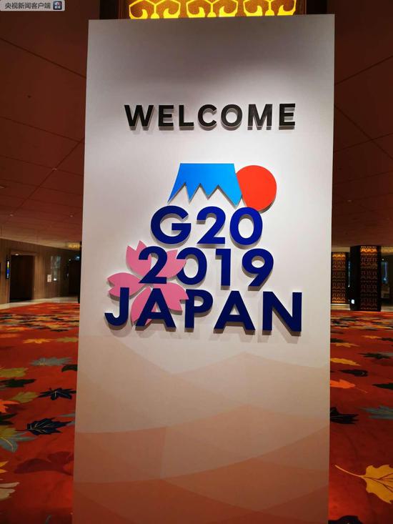 新闻中心的G20峰会标志牌。标志由象征着日本的富士山、太阳和樱花等元素构成。富士山的“山顶”和“朝日”象征着增长和繁荣。山脚下，盛开的樱花代表着春天，寄托了人们对全球经济繁荣增长的美好期待（央视记者荆伟拍摄）
