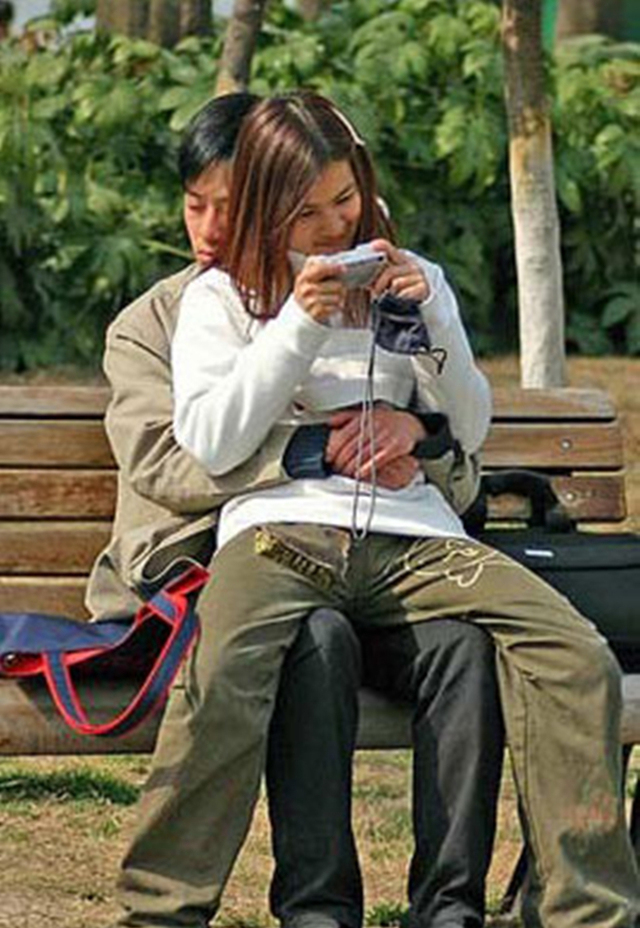 现代人在公园广场,女孩子坐在男孩子身上,一个打瞌睡,一个玩手机.