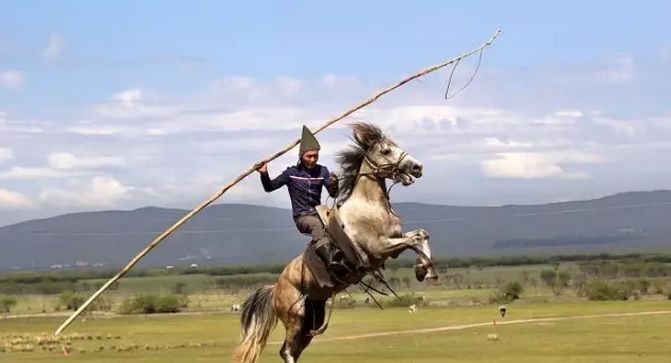 一位放牧人骑在马儿的身上,手上还拿着一根杆子,好像是在吓唬马