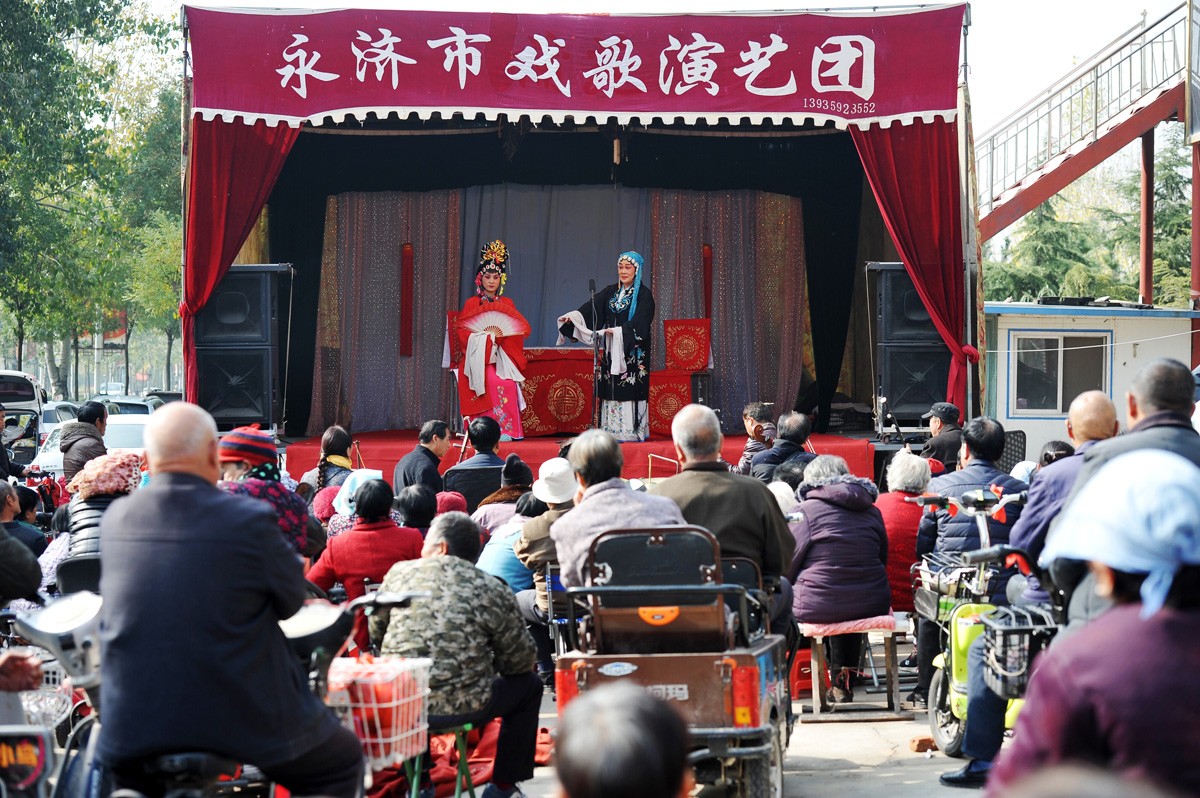 11月11日,山西永济,两名老人头顶遮挡阳光的纱巾在观看舞台上演出的